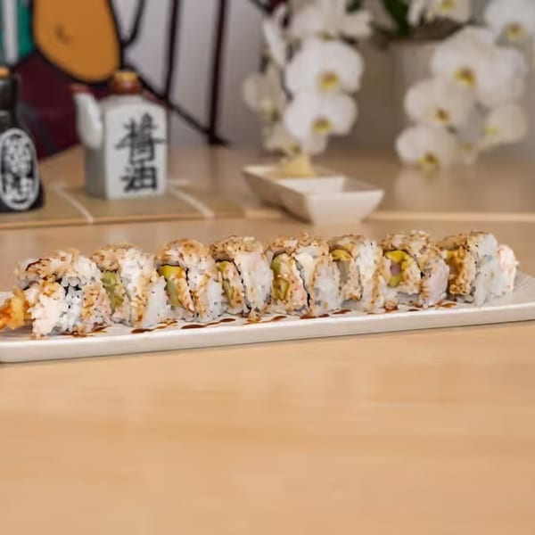 A long white plate of sushi rolls with shrimp tempura, drizzled with sauce, on a wooden table.