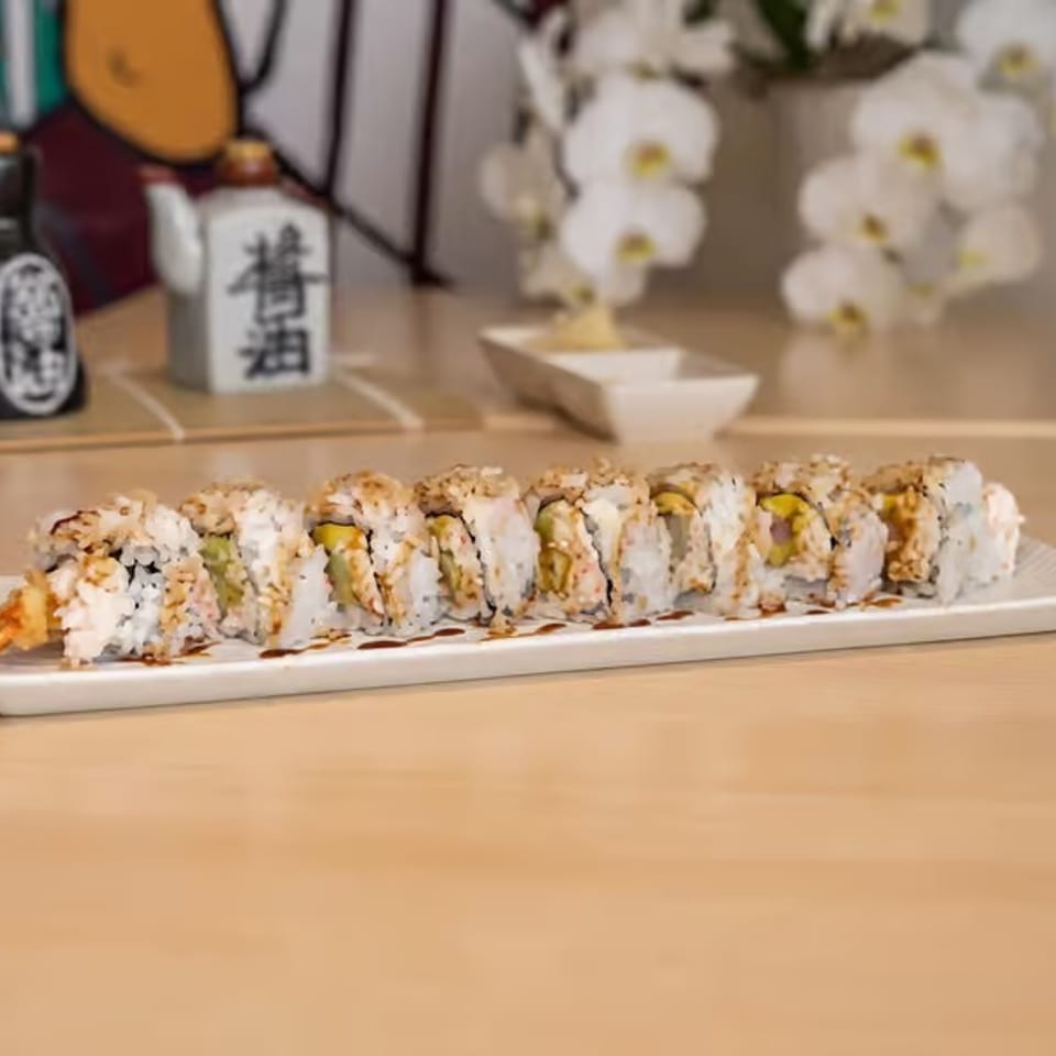 A long white plate of sushi rolls with shrimp tempura, drizzled with sauce, on a wooden table.