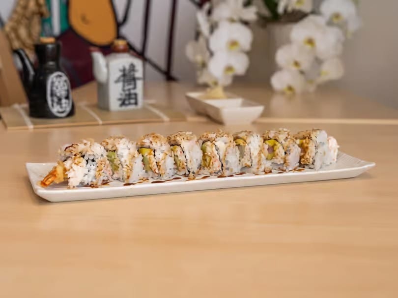 A long white plate of sushi rolls with shrimp tempura, drizzled with sauce, on a wooden table.
