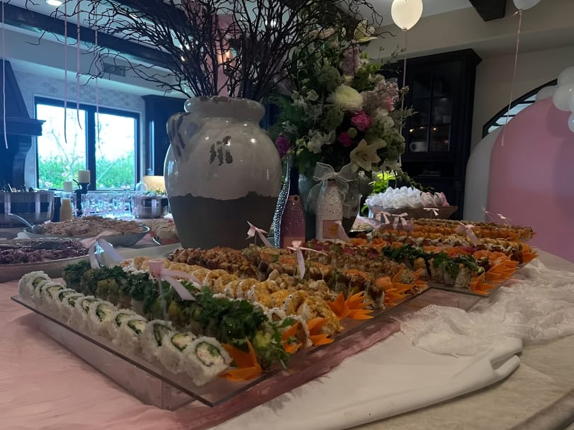 A buffet table laden with various sushi rolls, decorated with pink ribbons and floral arrangements.