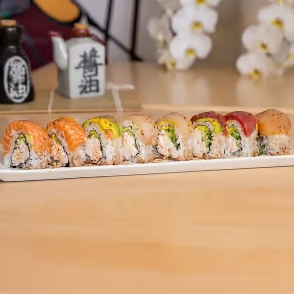 A white rectangular plate holds a row of colorful sushi rolls on a light wooden table.