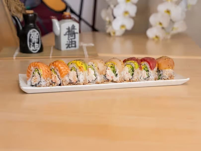 A white rectangular plate holds a row of colorful sushi rolls on a light wooden table.