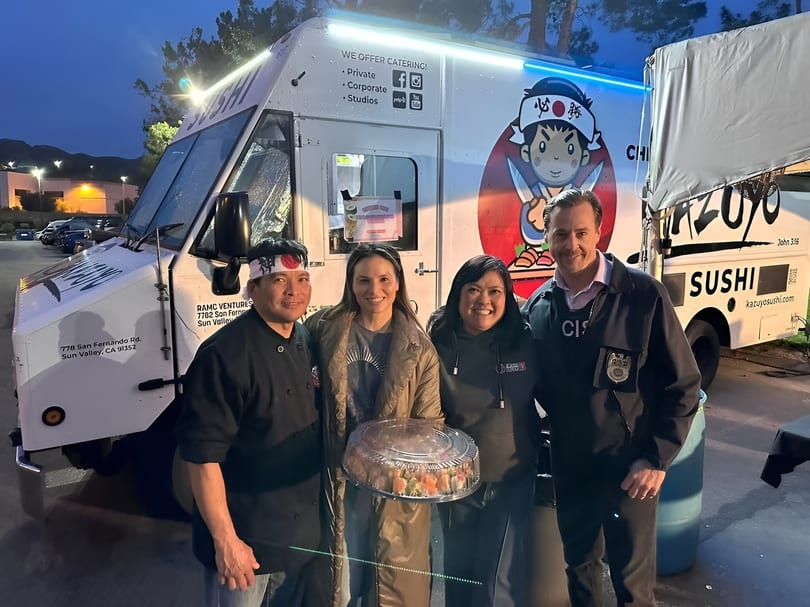 Four people, including a chef, pose with a large sushi platter in front of a sushi food truck.