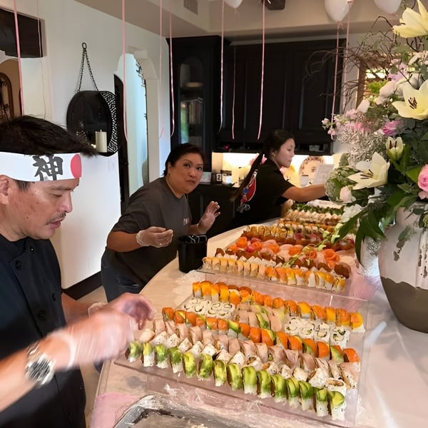 Chefs prepare a large sushi spread for a party, with balloons and flowers in the background.