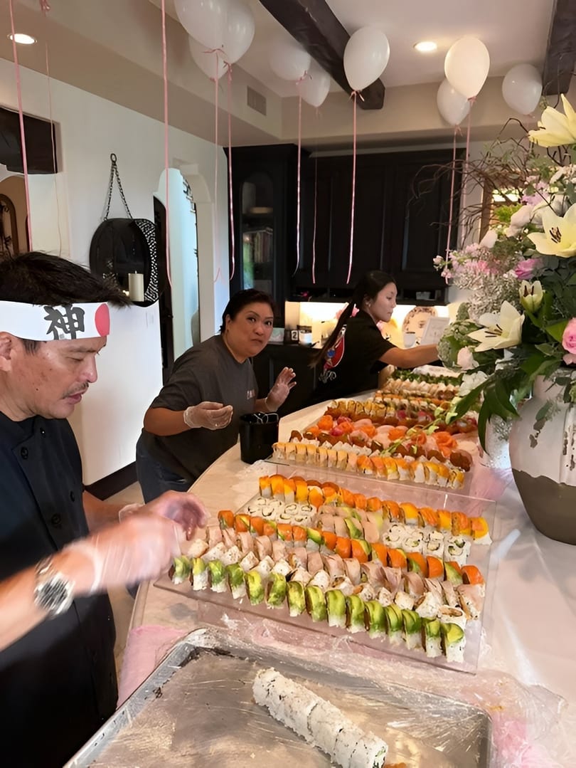 Chefs prepare a large sushi spread for a party, with balloons and flowers in the background.