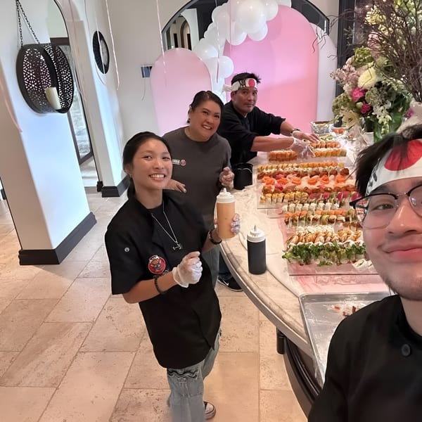 Three chefs smiling behind a sushi bar with many rolls, decorated with pink arches and balloons.
