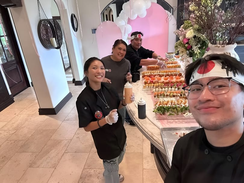 Three chefs smiling behind a sushi bar with many rolls, decorated with pink arches and balloons.