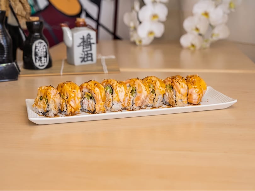 A white rectangular plate holds a row of sushi rolls topped with sauce and sesame seeds.